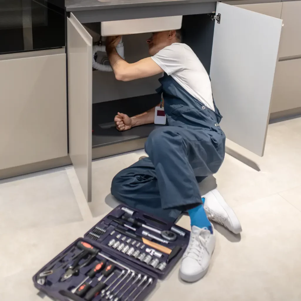 A person in a blue uniform set on the kitchen floor under cabinets to install the sink.