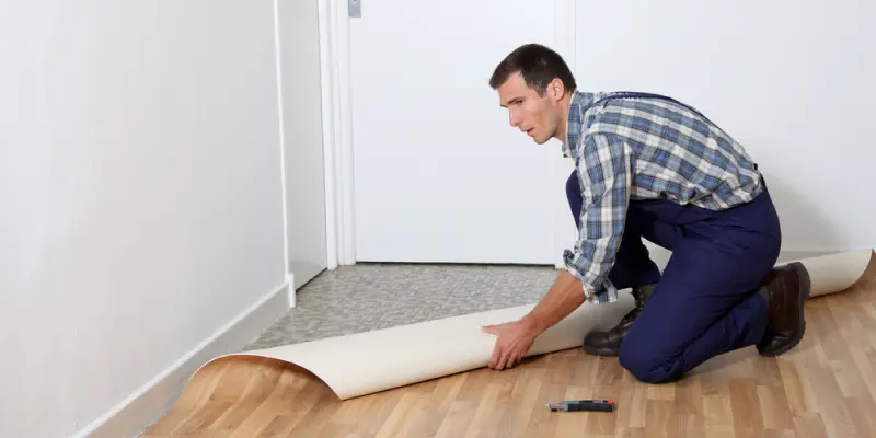 A handyman in blue pants and multi-color T-Shirt installing a vinyl sheet on the floor of a room